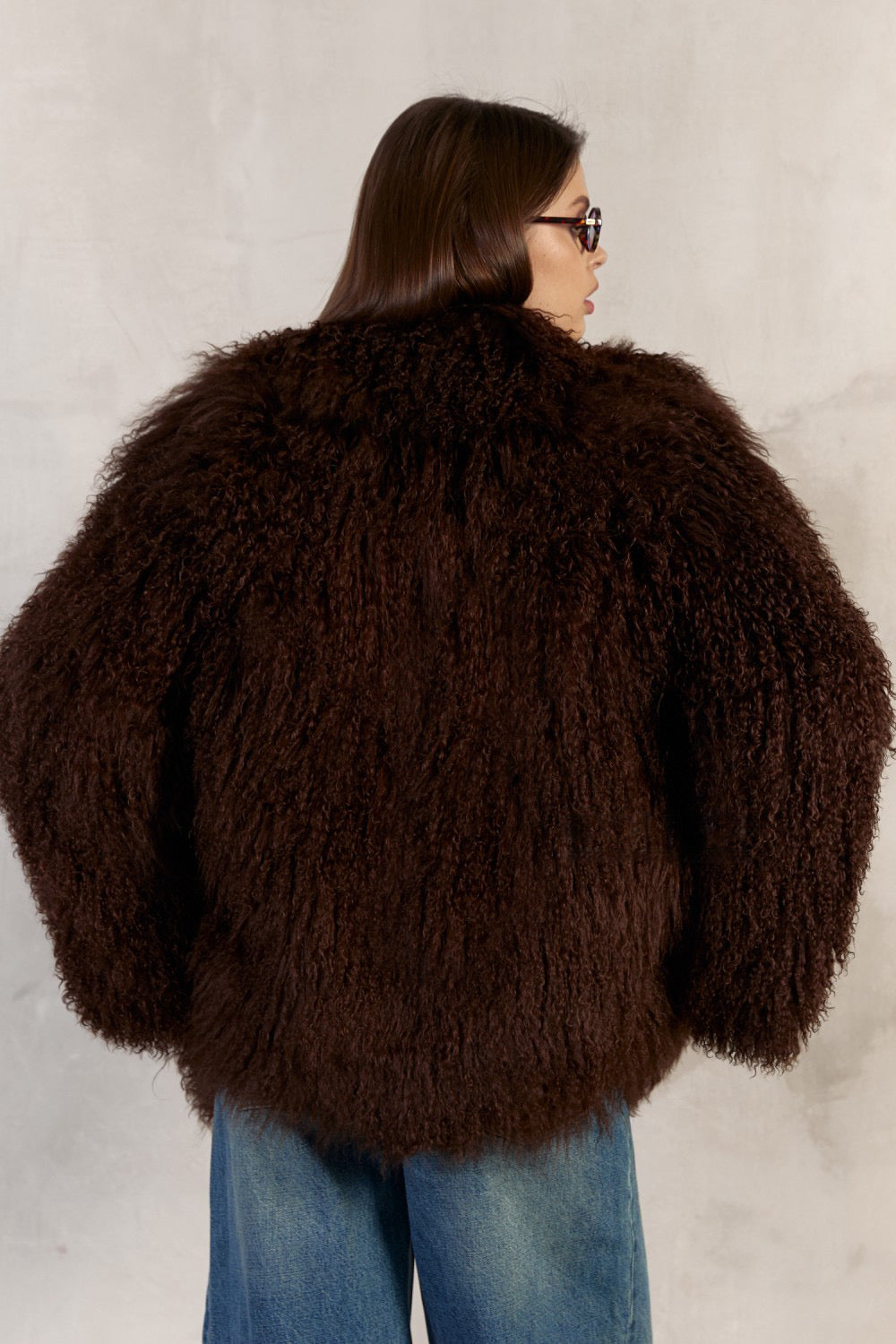 Wearing a genuine 100% Mongolian fur coat in rich brown, a woman exudes luxury with the coat’s dramatic collar, practical side pockets, and smooth satin and leather inner lining.
